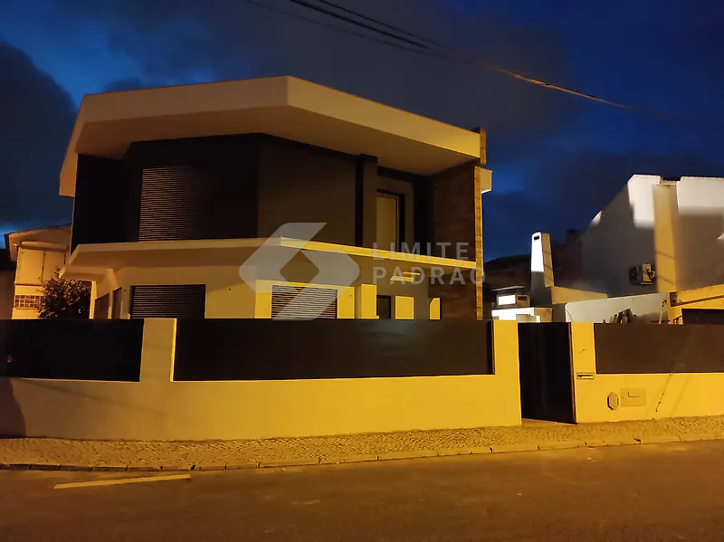 Construção de moradia: dois pisos, muro e portões, ao anoitecer