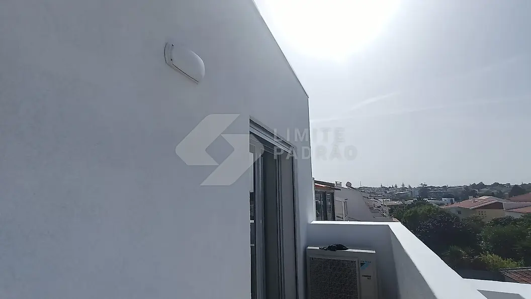 Pormenor de fachada: parede exterior rebocada e pintada de branco, com aplique e unidade exterior